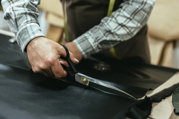 Partial view of male handbag craftsman cutting leather by scissors at studio — Stock Photo