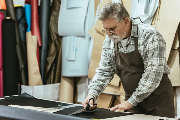 Зрелый мужчина ремесленник сумочки в фартуке и очки резки кожи ножницами в мастерской — стоковое фото