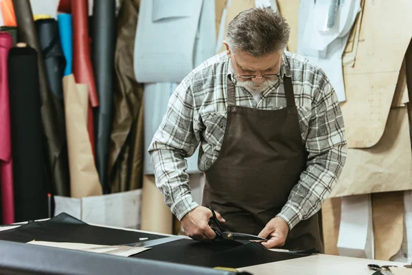 Мужская сумочка среднего возраста, мастер в фартуке и очках, режет кожу ножницами в мастерской — стоковое фото