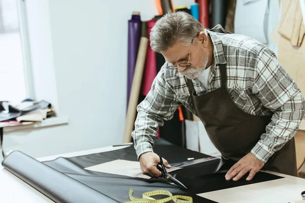 Male craftsman in apron and eyeglasses cutting leather by scissors at workshop — Stock Photo