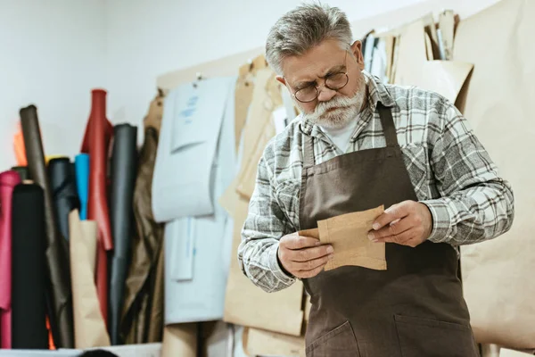 Artesano de bolsos de mediana edad enfocado en delantal y gafas que trabajan en el estudio - foto de stock