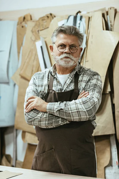 Cheerful middle aged craftsman in apron and eyeglasses standing with crossed arms at studio — Stock Photo