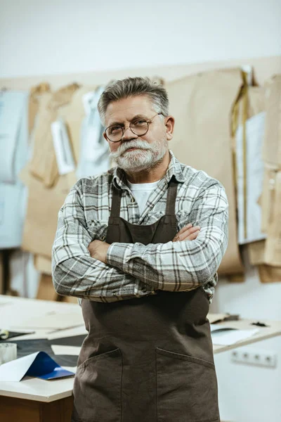 Lächelnder älterer Handwerker in Schürze und Brille steht mit verschränkten Armen im Atelier — Stockfoto