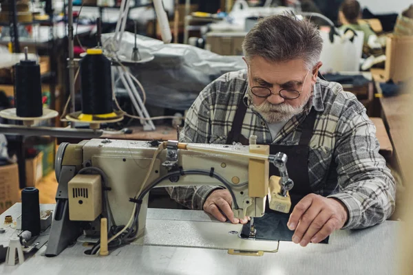 Sastre de mediana edad en delantal y anteojos que trabajan en la máquina de coser en el estudio - foto de stock