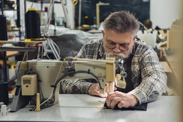 Fokussierte reife männliche Schneiderin in Schürze und Brille, die im Atelier an der Nähmaschine arbeitet — Stockfoto