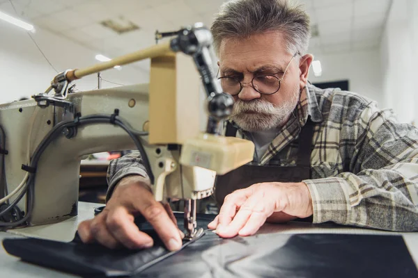 Enfoque selectivo de sastre macho maduro en delantal y anteojos que trabajan en la máquina de coser en el estudio - foto de stock