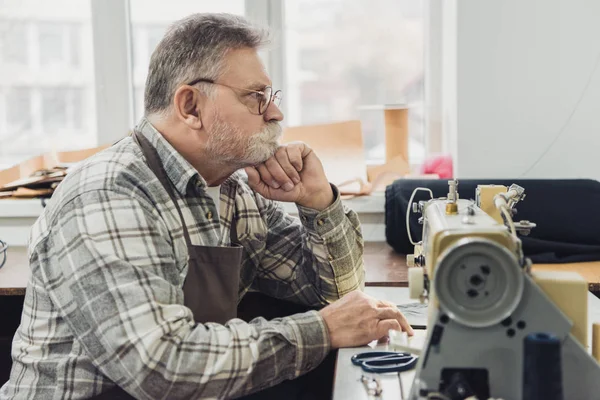 Side view of pensive mature male tailor in apron and eyeglasses sitting near sewing machine at studio — Stock Photo