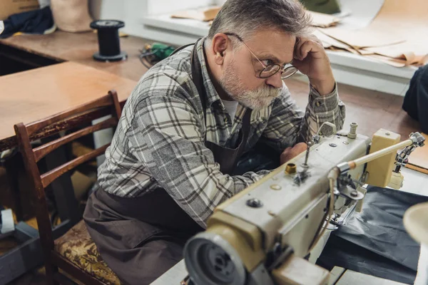 Vue grand angle du tailleur mâle mature dans le tablier et les lunettes assis près de la machine à coudre au studio — Photo de stock