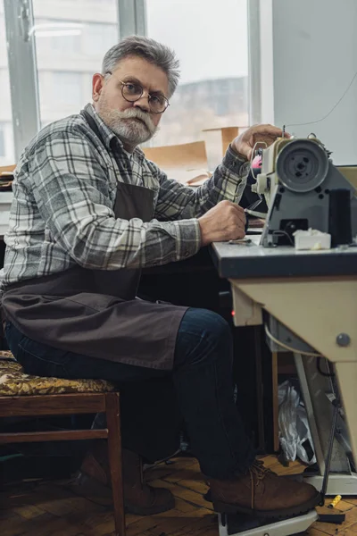 Sastre de mediana edad en delantal y anteojos posando cerca de la máquina de coser en el estudio - foto de stock