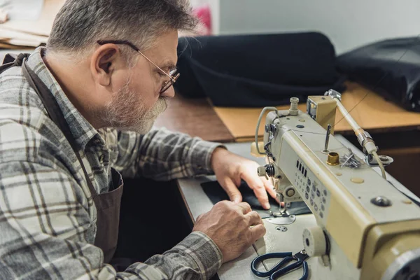 Vue latérale du tailleur mâle mature dans le tablier et les lunettes travaillant sur la machine à coudre au studio — Photo de stock