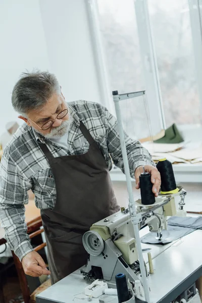 Focused middle aged tailor in apron setting strings on sewing machine at workshop — Stock Photo