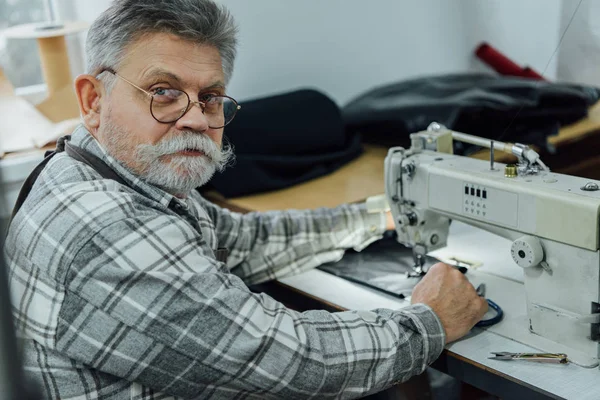 Portrait d'un tailleur homme mature dans un tablier et des lunettes assis près d'une machine à coudre en studio — Photo de stock