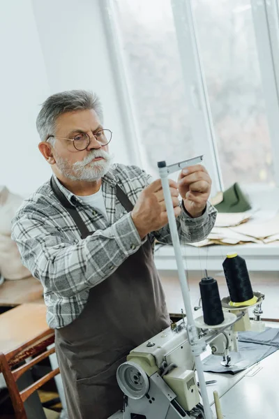 Focused middle aged tailor in apron cutting strings on sewing machine at workshop — Stock Photo