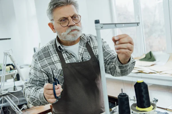 Focused mature male tailor in apron setting strings on sewing machine at workshop — Stock Photo