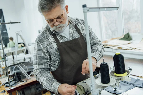 Selective focus of mature male tailor in apron setting strings on sewing machine at workshop — Stock Photo