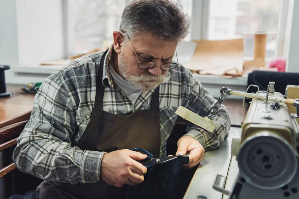 Reifer männlicher Schneider in Brille und Schürze schneidet Leder mit der Schere in der Werkstatt — Stockfoto