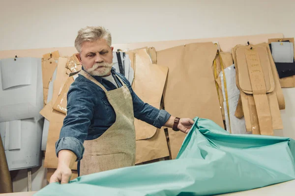 Messa a fuoco selettiva di mezza età artigiano borsa maschile mettendo tessuto sul tavolo di lavoro in studio — Foto stock