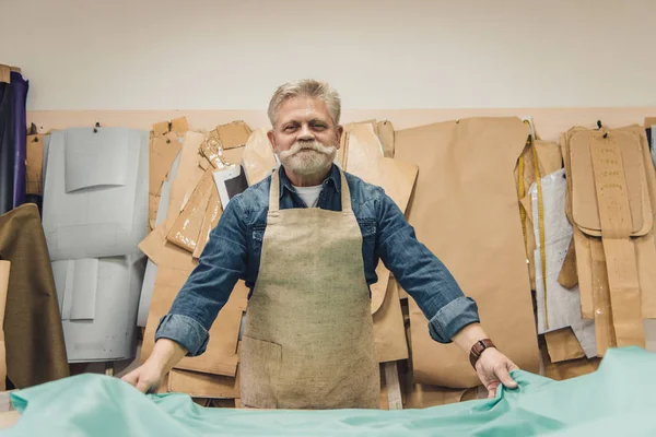Alegre maduro masculino bolsa artesão colocando tecido na mesa de trabalho no estúdio — Fotografia de Stock