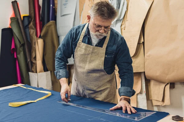 Зосереджений чоловічий майстер середнього віку в фартуху робить вимірювання на тканині в майстерні — стокове фото