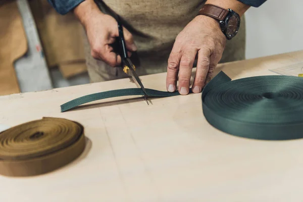 Partial view of male handbag craftsman cutting fabric by scissors at workshop — Stock Photo