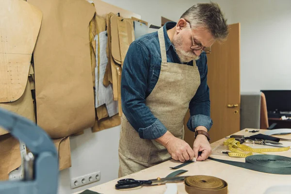 Selective focus of middle aged male handbag craftsman in apron and eyeglasses working with fabric at studio — Stock Photo