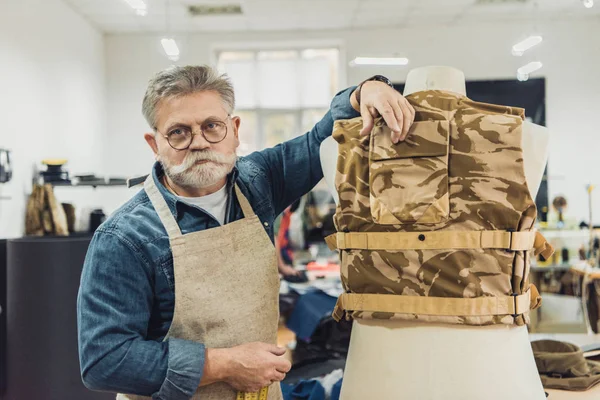 Alegre sastre masculino de mediana edad de pie cerca de chaleco militar en el estudio - foto de stock