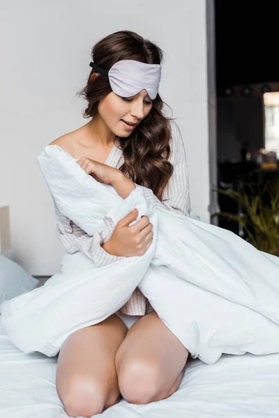 Нежная брюнетка девушка в маске для сна сидит на кровати с одеялом по утрам — стоковое фото