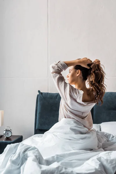 Brunette young woman making pony tail while sitting on bed in the morning — Stock Photo