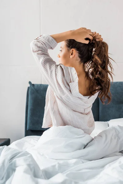 Brunette woman making pony tail while sitting on bed in the morning — Stock Photo