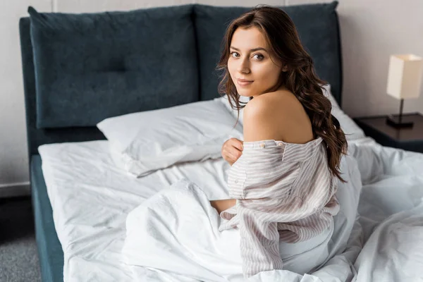 Улыбающаяся нежная девушка в пижаме, сидящая на кровати по утрам — стоковое фото