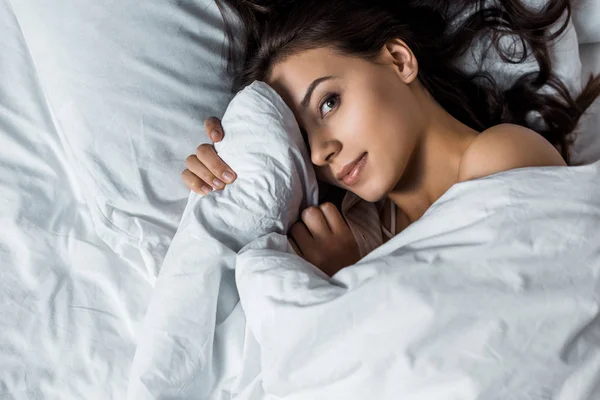 Brunette girl looking at camera and hiding under white blanket on bed — Stock Photo