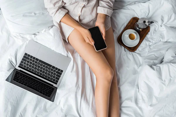 Vista ritagliata di donna in possesso di smartphone mentre seduto sul letto con moka caffè e laptop — Foto stock