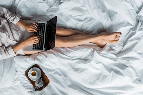 Vista sezione bassa della donna con tazza di caffè e moka pot utilizzando il computer portatile a letto — Foto stock