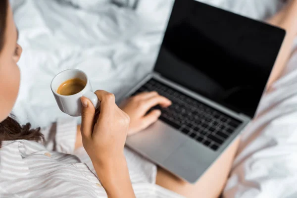 Cropped view of girl with coffee using laptop with blank screen in bed — Stock Photo