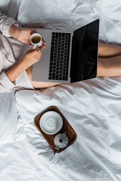 Vista cortada da mulher com xícara de café e moka pot usando laptop na cama — Fotografia de Stock