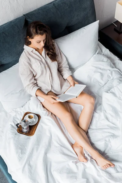 Hermosa chica leyendo libro en la cama con cafetera italiana por la mañana - foto de stock