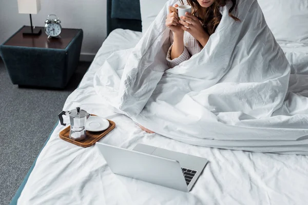 Vista cortada da mulher com xícara de café olhando para laptop na cama — Fotografia de Stock