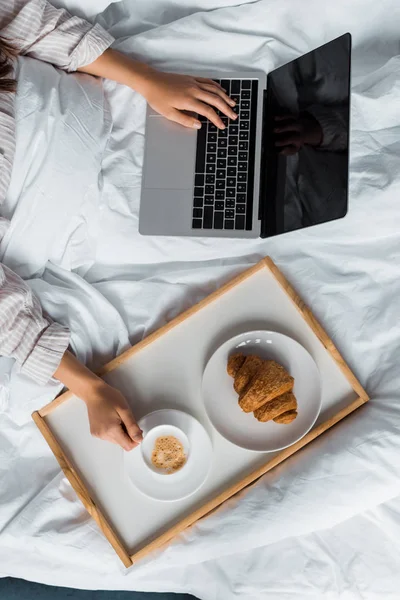 Vista ritagliata di donna con computer portatile che fa colazione con croissant e caffè a letto — Foto stock