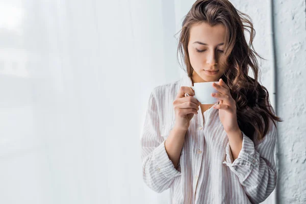 Junge brünette Frau, die morgens mit einer Tasse Kaffee am Fenster steht — Stockfoto