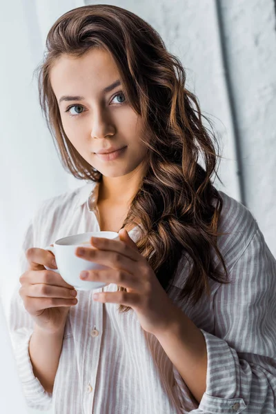Красива молода жінка стоїть з чашкою кави біля вікна і дивиться на камеру — стокове фото