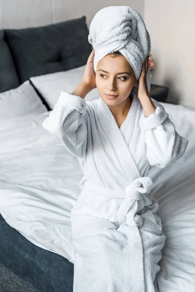 Schönes Mädchen im weißen Bademantel, das Handtuch auf dem Kopf trägt, während es auf dem Bett sitzt — Stockfoto