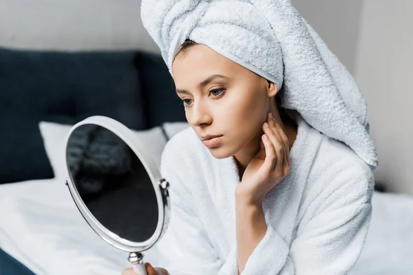 Beautiful woman in white bathrobe and towel looking at her skin in mirror — Stock Photo