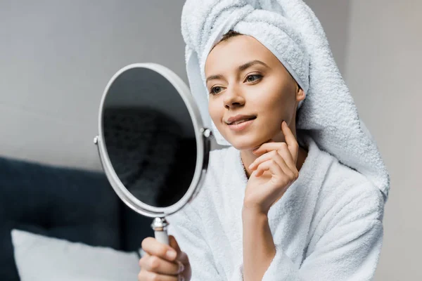 Happy woman in white bathrobe and towel looking at her clean face in mirror — Stock Photo