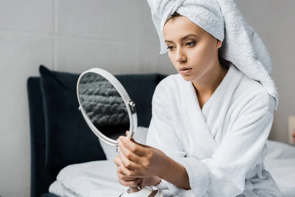 Attractive girl in white bathrobe and towel looking at her face in mirror — Stock Photo