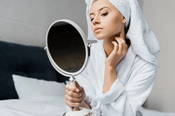 Young woman in white bathrobe and towel looking at her skin in mirror — Stock Photo