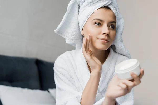 Attraktive junge Frau in Bademantel und Handtuch auf dem Kopf, die Feuchtigkeitscreme auf das Gesicht aufträgt — Stockfoto