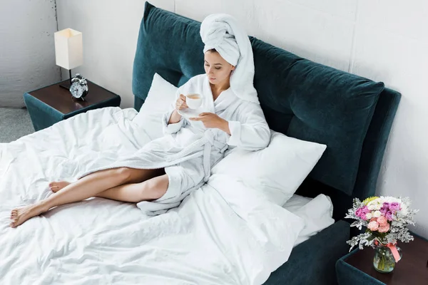 Hermosa chica en albornoz y toalla blanca sosteniendo taza de café mientras descansa en la cama por la mañana - foto de stock