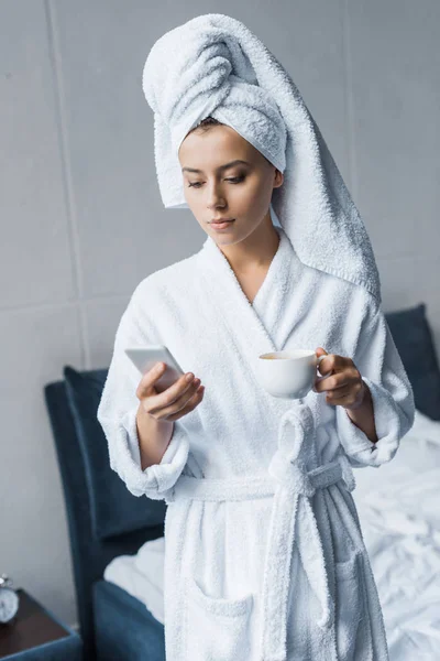 Young woman in white bathrobe and towel holding cup of coffee and using smartphone in bedroom — Stock Photo