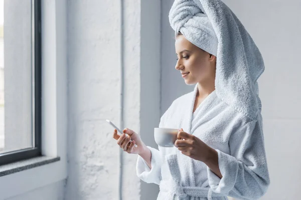 Atractiva mujer joven en albornoz sosteniendo la taza de café y el uso de teléfono inteligente cerca de la ventana - foto de stock