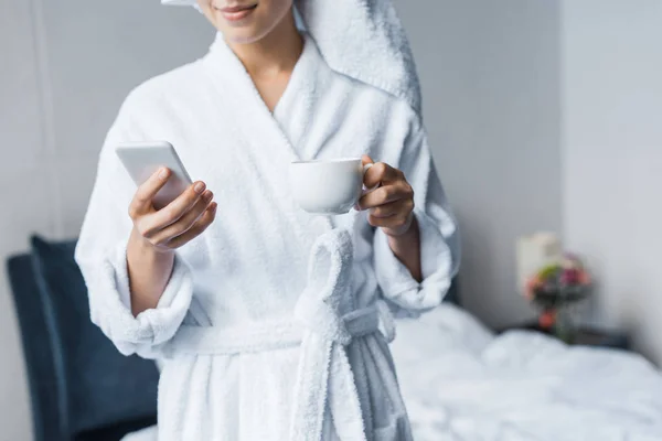 Vista recortada de la mujer en albornoz sosteniendo taza de café y el uso de teléfono inteligente en el dormitorio por la mañana - foto de stock